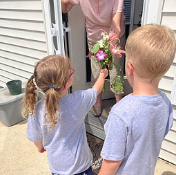 Two small kids hand a bouquet of flowers to an elderly woman on her doorstep