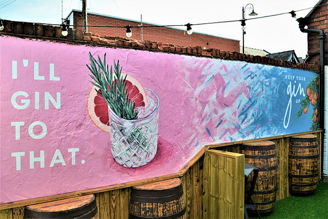 Outdoor hand-painted mural in a pub gin garden with a colourful pink and blue background, a glass of gin, and text saying "I'll gin to that"