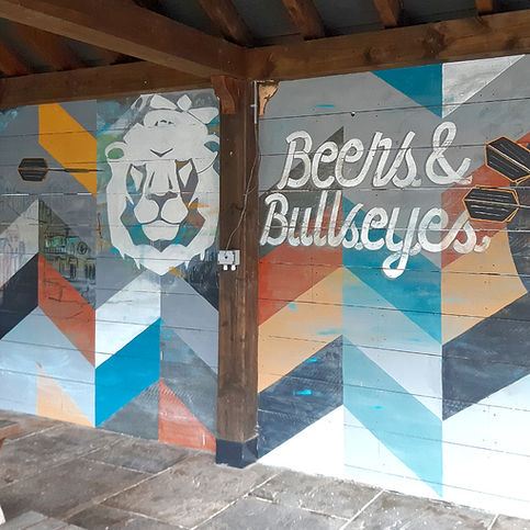 A long outdoor mural along a covered seating area, the piece was designed with designers from the sub-contracting company. The venue was a concept pub where people could play a variety of electronic darts games.
The mural incorporated elements of the darts theme, the 'White Lion' pub name and branding, as well as the Macclesfield location. An abstracted landscape of the town center was included in the middle, to the left side a darts player wearing a lions mane throws a dart across the entire mural towards the dart board on the right hand side.