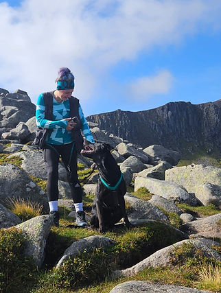 Standing on hilly terrain, with some large boulders a girl is stood next to her black dog as they both look at each other.