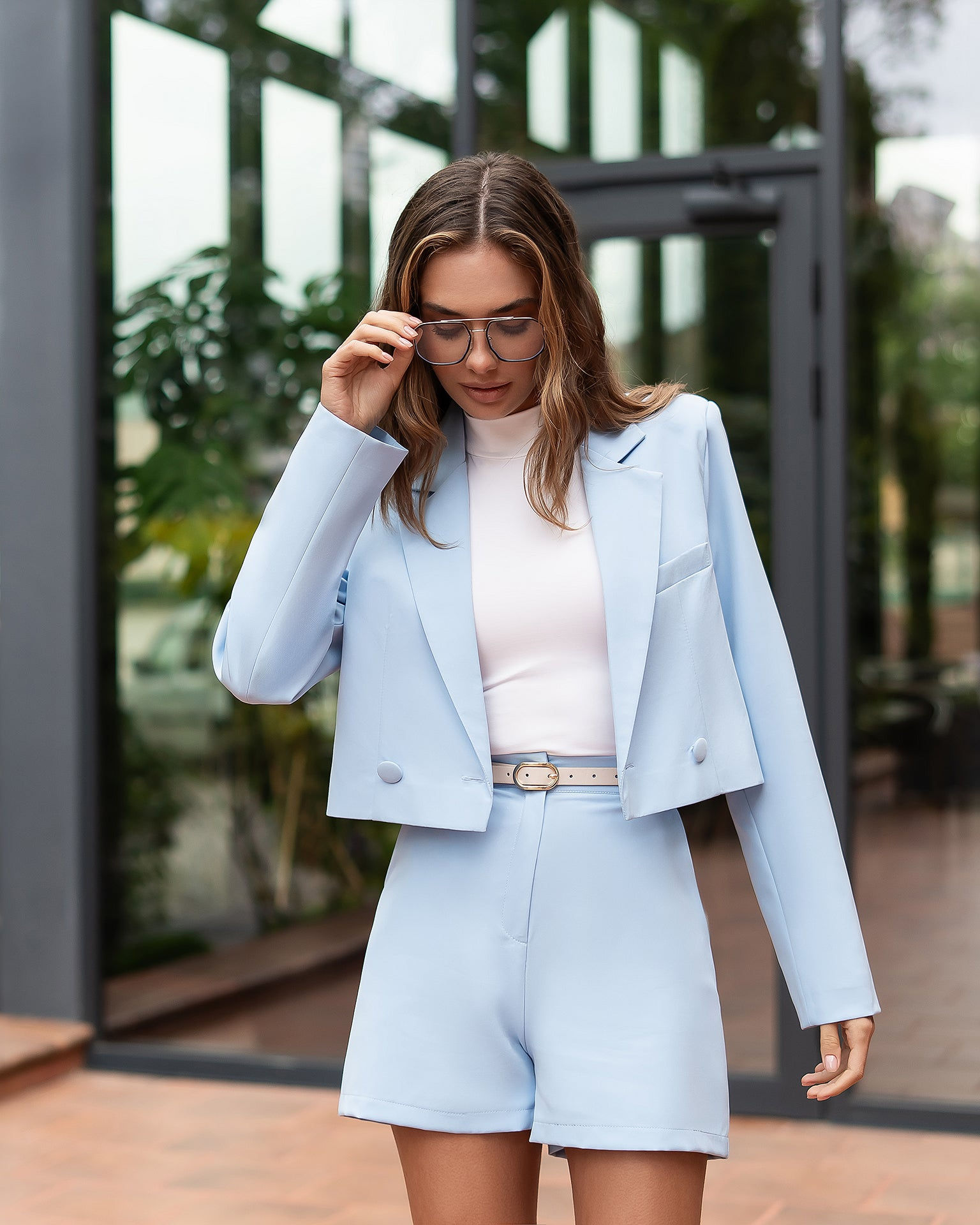 Joven profesional ajusta sus gafas, traje azul claro, exterior oficina.