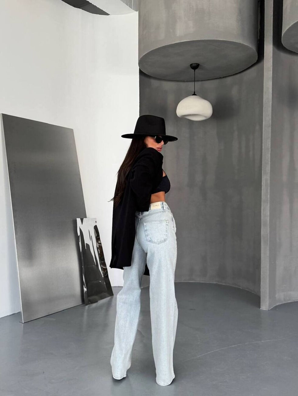 Miniatura: Mujer de espaldas con sombrero negro, jeans, en habitación moderna.