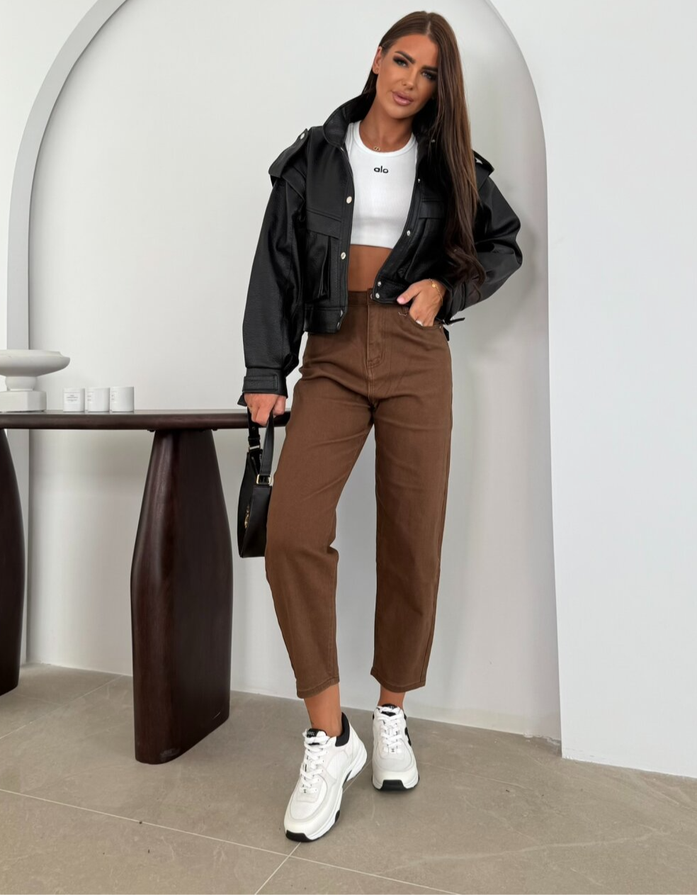 Mujer vestida con chaqueta de cuero negra, top blanco y pantalones marrones.