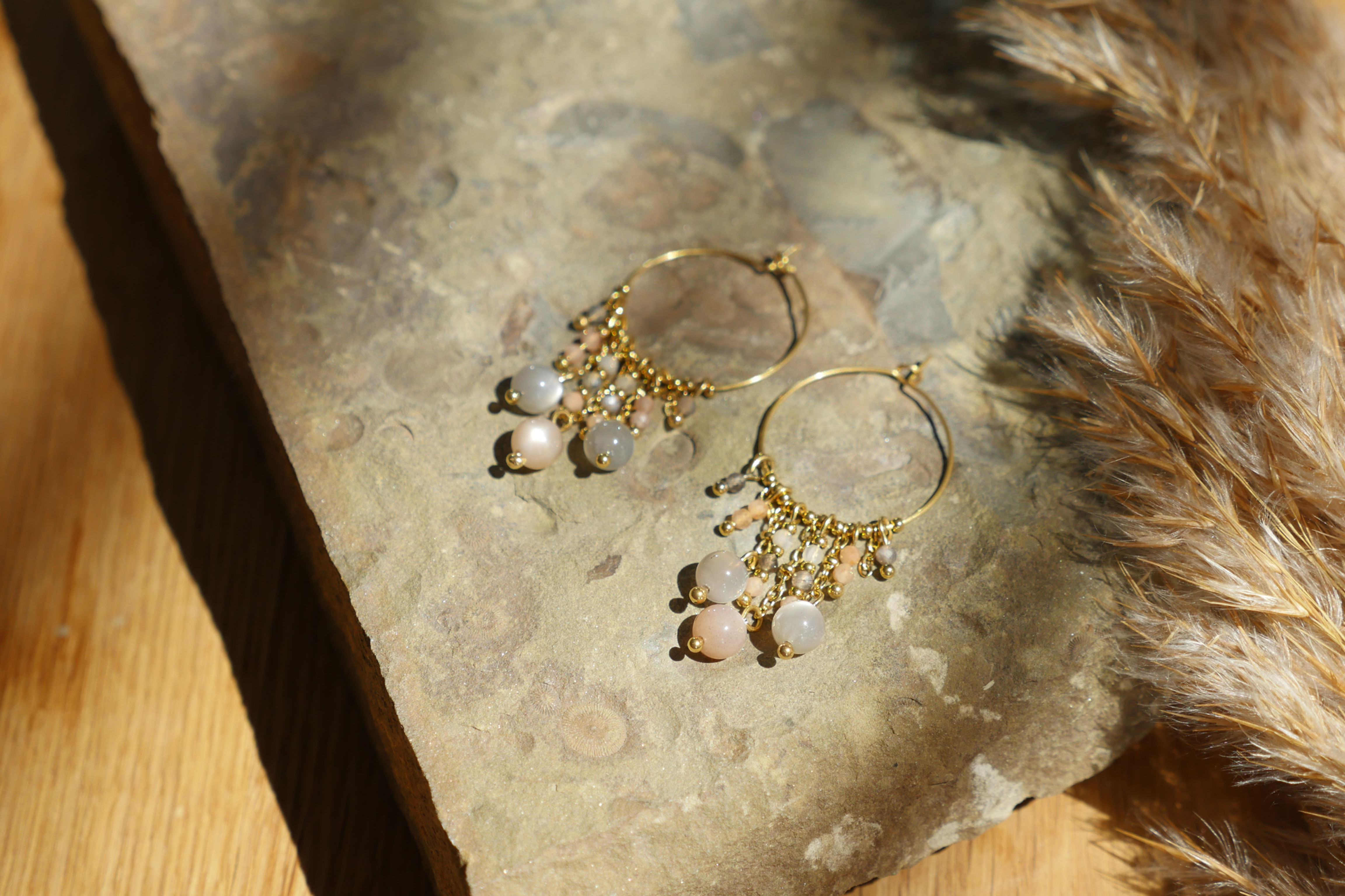 Moon Whisper - Creolen aus vergoldetem Edelstahl mit Mondstein auf Naturstein mit Pampasgras im Hintergrund.