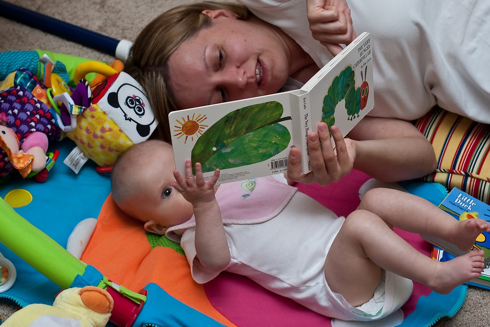 A baby wearing a white onsie and pink bib lays on a colorful mat surrounded by toys. A woman with blond hair and a white tshirt lays next to the baby as they both hold The Very Hungry Caterpillar book. Woman is pointing to a page.
