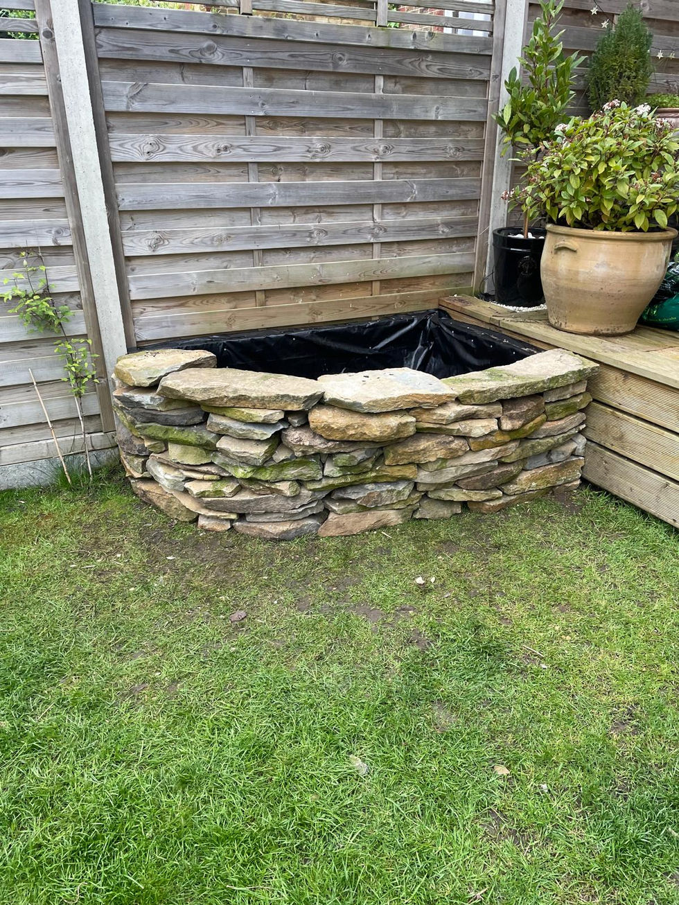 Dry-stone corner planter built from stacked natural stone, planted with an olive tree and low shrubs against a timber fence.