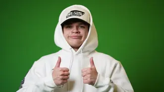 Jynxzi smiling and giving two thumbs up wearing a white hoodie and cap against a green background