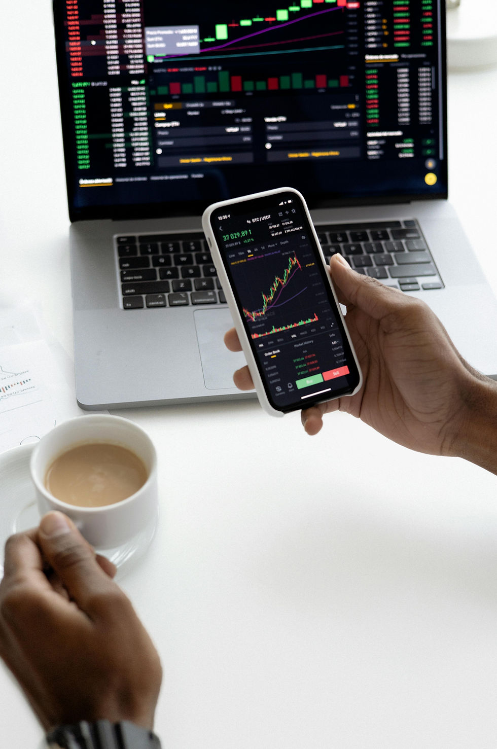 Hands hold a smartphone with a stock chart, in front of a laptop displaying similar graphs. A cup of coffee sits nearby on a white table.