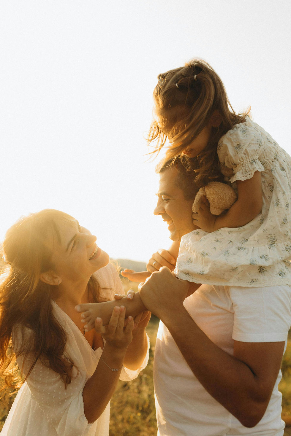 Smiling family in a sunny field. Man carries a girl with a teddy on his shoulders, woman holds girl's foot, all in warm tones.