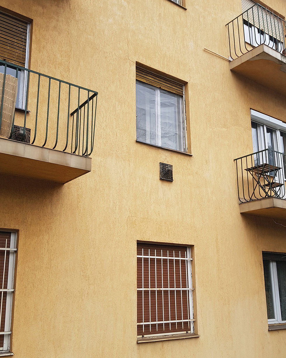 Sárga vakolatú társasház homlokzata erkélyekkel, zárt redőnyös ablakokkal és zöld/fekete korlátokkal. Az ablak alatt konvektor parapet.