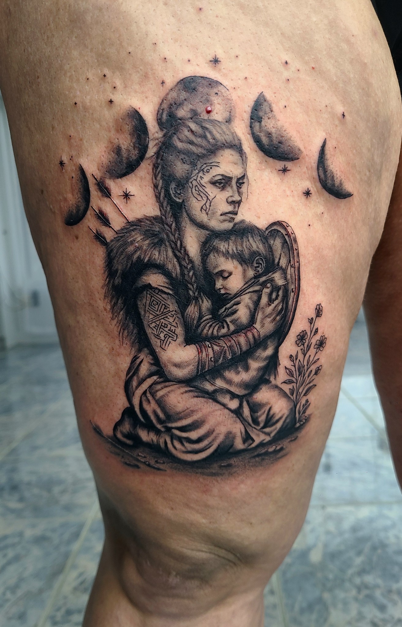 Black-and-grey thigh tattoo of a seated figure holding a child, surrounded by crescent moon phases