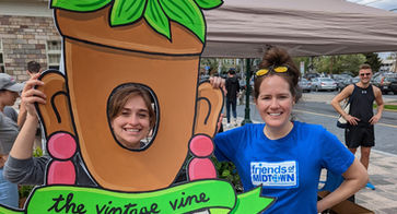 Two women with one woman putting her face through a cutout in a big paper plant with "The Vintage Vine" on it
