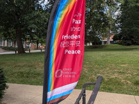 A tall feather flag outside Grace Episcopal Church displays the word “Peace” in multiple languages, including Arabic, Spanish, Hebrew, German, Chinese, and English. The flag has a rainbow design across the top and a rainbow-striped border at the bottom. The church’s name and logo appear near the base of the flag. The background shows a grassy lawn, sidewalk, and trees.