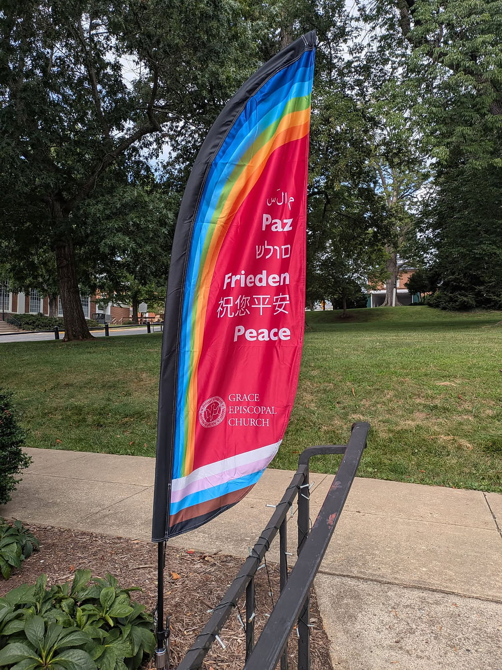 A tall feather flag outside Grace Episcopal Church displays the word “Peace” in multiple languages, including Arabic, Spanish, Hebrew, German, Chinese, and English. The flag has a rainbow design across the top and a rainbow-striped border at the bottom. The church’s name and logo appear near the base of the flag. The background shows a grassy lawn, sidewalk, and trees.