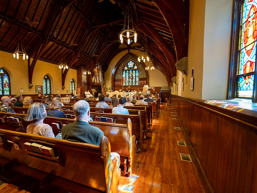 The image shows the interior of a traditional church during a service. The church has wooden pews with people seated, facing the altar at the front. The interior features high, vaulted wooden ceilings with exposed beams and stained glass windows lining the walls. The lighting is warm, provided by hanging chandeliers, and there is a sense of calm and reverence in the atmosphere.