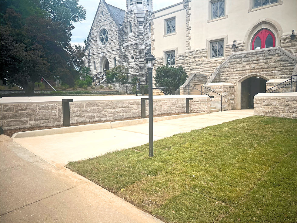 Stone church buildings with arched entrances, red doors, stairs, and a newly paved accessible walkway bordered by low stone walls, black railings, lamp posts, and a grassy lawn.