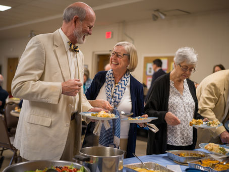 Three older adults serve themselves from a buffet line at a community meal. A man in a cream-colored suit and bow tie smiles warmly at a woman beside him wearing glasses and a blue and white scarf. Another woman with short white hair and glasses, dressed in a patterned blouse and black cardigan, focuses on selecting food. The setting appears to be a cheerful gathering in a church or community hall, with trays of food and a pot of salad in the foreground.