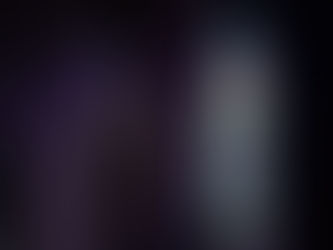 A dimly lit church interior showing two decorative metal crosses illuminated by colorful light from a tall stained glass window. The stained glass features geometric patterns in red, blue, yellow, and white. A small bookshelf with pamphlets is partially visible on the left side of the image.