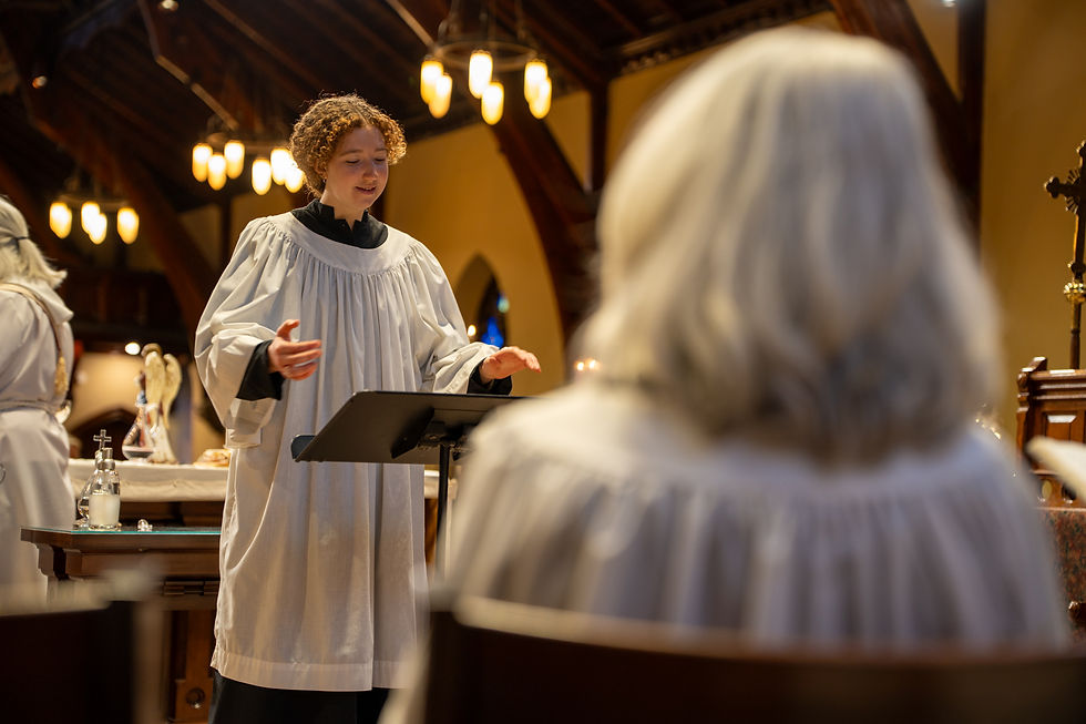 A young acolyte in a white robe stands at a lectern inside a warmly lit church, gesturing as they speak or read. Wooden beams and hanging lights line the ceiling, and candles and altar items are visible behind them. In the foreground, slightly out of focus, another robed person sits facing the speaker.