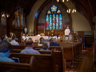 A congregation sits in wooden pews during a church service as a priest speaks from the pulpit near the altar, framed by stained-glass windows, organ pipes, and arched wooden beams.