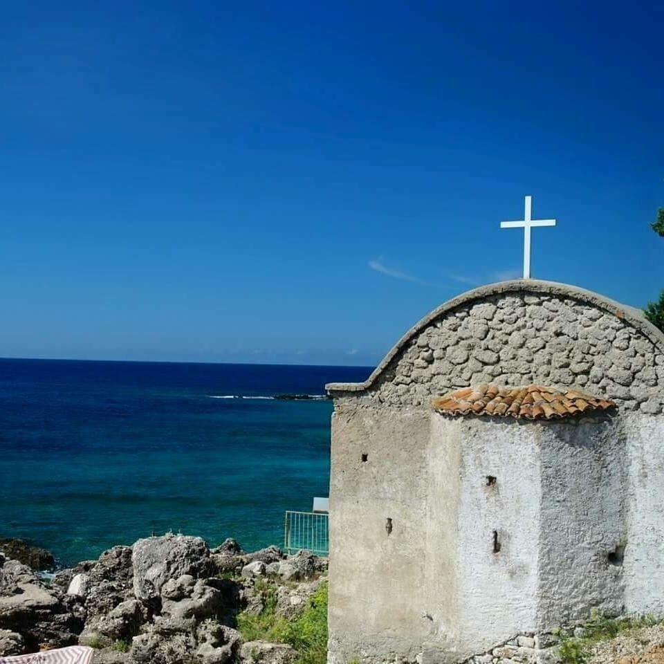 Kishë e vogël me çati qeramike dhe kryq, pranë detit të kaltër nën qiell të kthjellët. Atmosferë paqësore bregdetare.