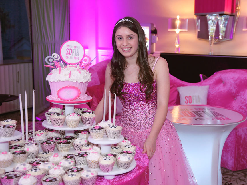 An excited young woman stands in front of a table filled with cupcakes at her Bat Mitzvah
