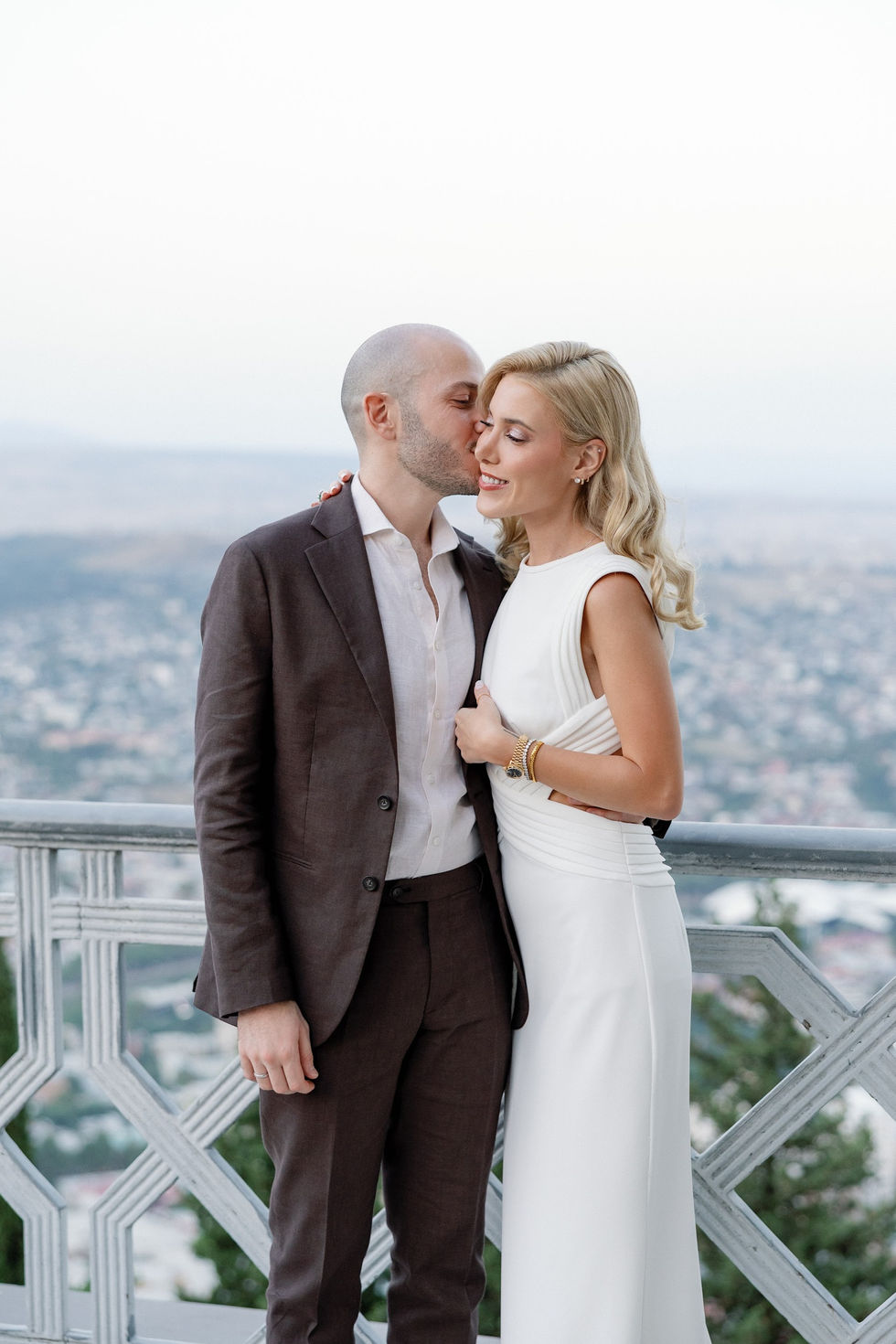 Romantic couple sharing a quiet moment on a scenic terrace overlooking Tbilisi, with soft daylight, elegant attire, and panoramic city views creating an intimate, timeless atmosphere.