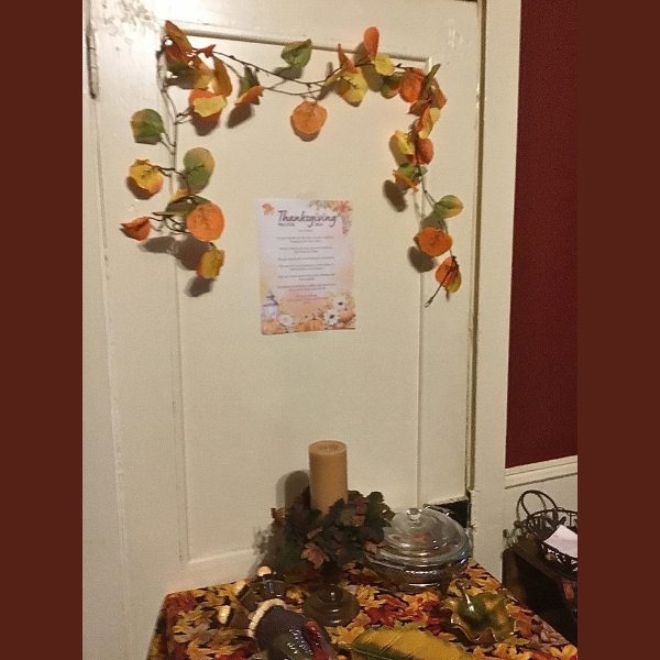 A cozy Thanksgiving nook adorned with autumn leaves and a themed table setting, featuring a festive candle centerpiece and decorative prayer on the door.