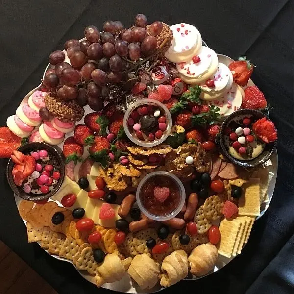 Colorful platter with grapes, strawberries, crackers, cheese, mini sausages, cookies, and dips on a black tablecloth. Festive and inviting.