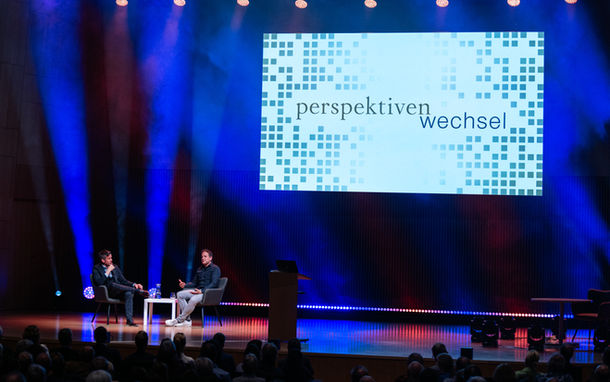 Der Moderator und der Referent sitzen auf der Bühne vor der Leinwand