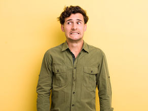 Young professional in an olive green shirt on a yellow background, looking anxious and struggling with self-doubt due imposter syndrome.