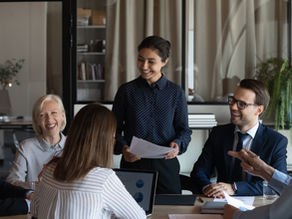 Diverse group of five business professionals seated around a table at a meeting with another coworker standing at the table.