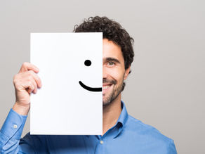 A business professional in a button-down shirt seen smiling and covering the other half of his face with a paper smiley face.