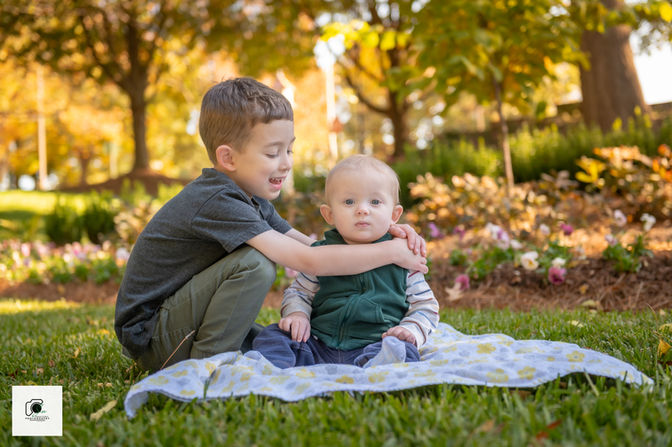 Older brother hugging his baby sibling on a flowered blanket in a park with golden autumn leaves in the background