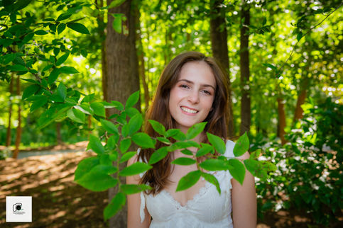 Young woman smiling behind leafy green branches in a sunlit forest — Novitsky Photography