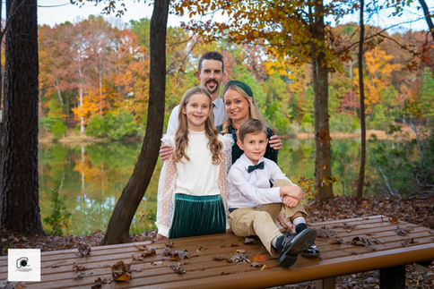 A family of four poses on a wooden bench near a lake with vibrant autumn trees in the background.
