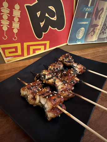 Grilled unagi skewers with sesame seeds on black plate, visible "肉" character.