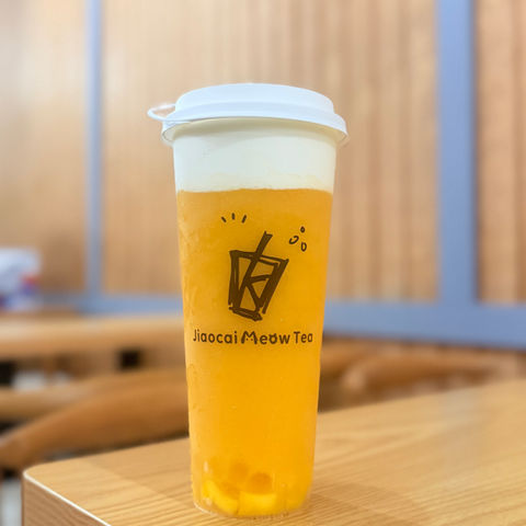 橙黄色奶盖茶，杯身印有“Maocai Meow Tea”，置于木桌。