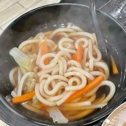 Steaming bowl of udon noodles with carrots and vegetables in broth.