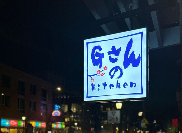 Lit restaurant sign for Gさんの kitchen with cherry blossoms at night.
