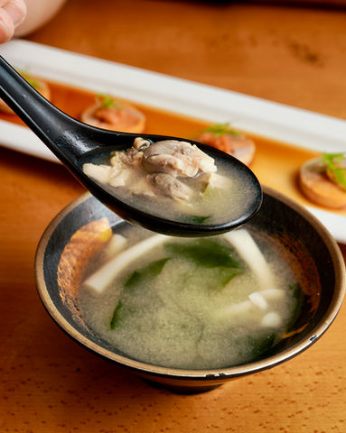 Spoon holding soup with meat, bowl of soup, and Yoi Sushi on the table.