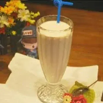Tall glass of milkshake with straw on table near flowers.