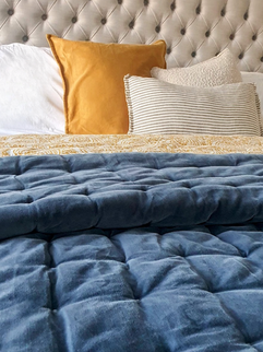 A navy velvet quilted throw over a double bed with scatter cushions in neutral tones and a pop of yellow
