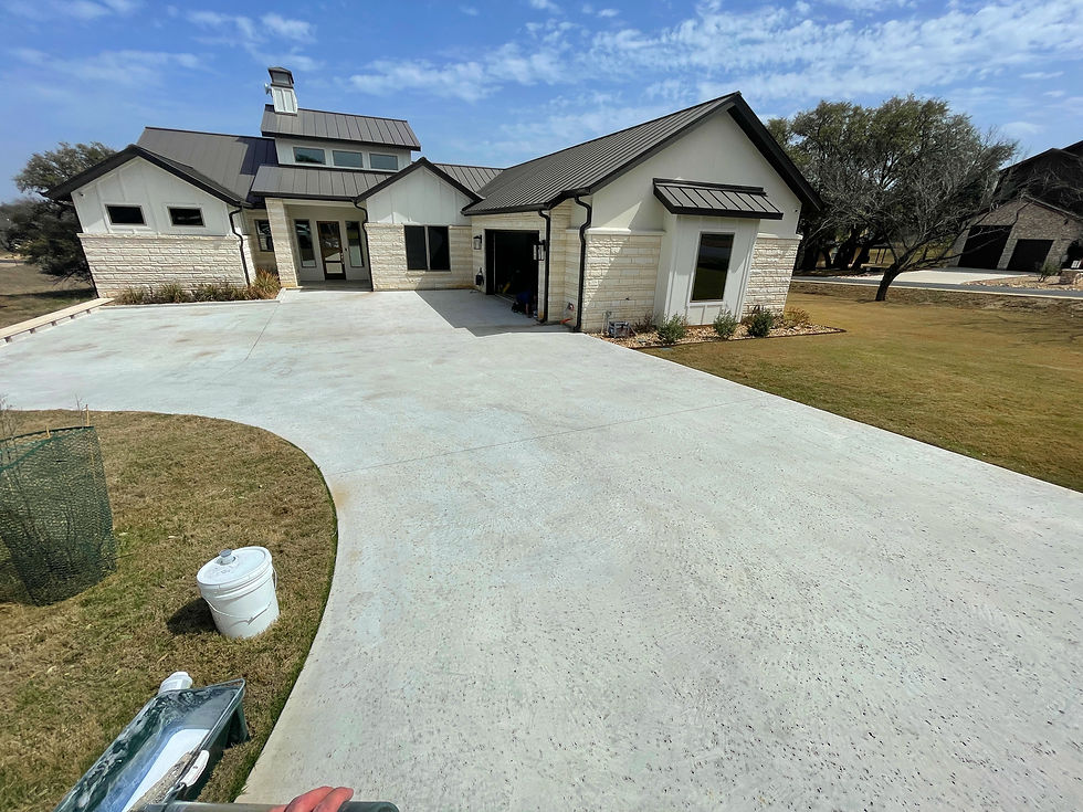 A wide-angle, ground-level photograph capturing a weathered, gray concrete residential driveway in Austin, Texas, showing significant neglect. Multiple prominent, heavy black hot tire marks and dark plasticizer migration streaks mar the surface near the center, set against the rough, porous substrate. Diffuse oil stains, localized dirt accumulation, and traces of greenish-black organic growth near the edges are visible, emphasizing a dull and worn appearance. The background shows an unkempt lawn and the lower section of a brick home under bright, harsh sunlight, highlighting the lack of surface protection.