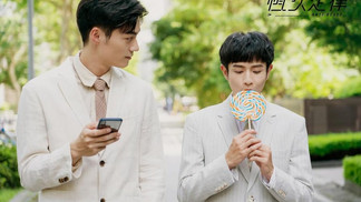 Two men walking outside, one holding a large lollipop and the other looking at a phone, suggesting a casual and playful moment.