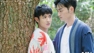 Two men standing close together in a forest, one leaning against a tree and the other looking at him intently, suggesting a tender and intimate moment.