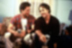 Two men are sitting together on a couch, smiling and laughing while holding cans of beer. One man is wearing a red plaid shirt over a white t-shirt, and the other is dressed in a dark shirt. The casual, friendly atmosphere suggests a moment of camaraderie and connection between the two. The scene appears warm and lighthearted, capturing a genuine and relaxed interaction. This image is likely from the movie The Sum of Us, which explores relationships and family dynamics.