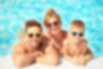 Mann, Frau und ein Kind liegen im Wasser und stützen sich am Beckenrand ab. Alle drei haben eine Sonnenbrille auf und lächeln.