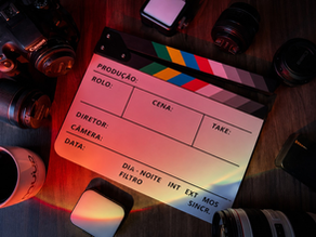 Mesa de madeira escura com equipamentos de produção audiovisual organizados ao redor de uma claquete de cinema posicionada no centro. A claquete está em branco, com campos para produção, rolo, cena, diretor, câmera, data e take. Ao redor, há uma câmera fotográfica, diversas lentes, um smartphone, um pequeno dispositivo eletrônico e uma caneca de café. A iluminação em tons vermelhos, azuis e alaranjados cria um efeito cinematográfico, com reflexos coloridos sobre a claquete e os objetos, transmitindo um ambiente de gravação e produção de conteúdo profissional.