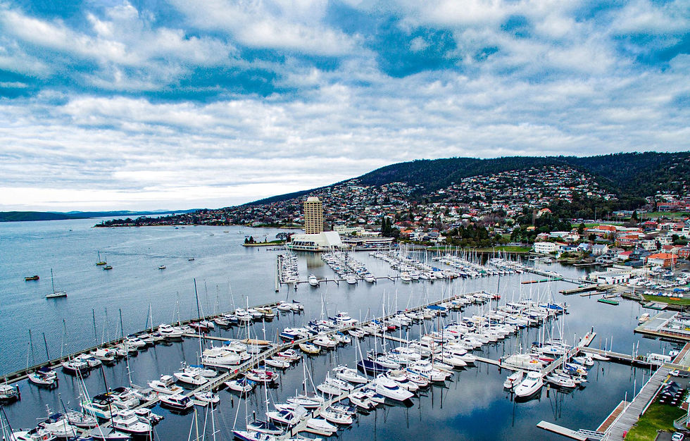 Aerial view of a marina packed with sailboats and yachts beside a hillside Sandy Bay under a cloudy blue sky.